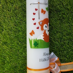 May include: White candle decorated with a cross, red hearts, and orange butterflies. A glittery orange fox is featured on the candle, along with the text "Zur Taufe" and a name and date. An orange ribbon is tied around the base.