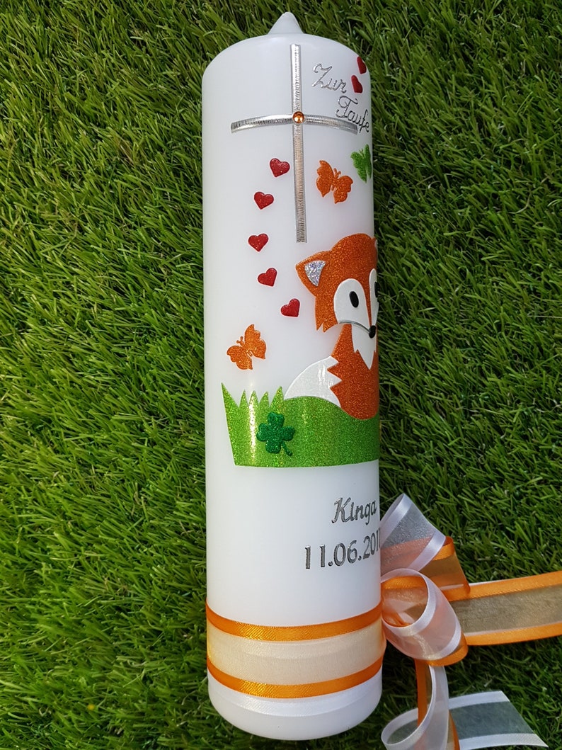 May include: White candle decorated with a cross, red hearts, and orange butterflies. A glittery orange fox is featured on the candle, along with the text "Zur Taufe" and a name and date. An orange ribbon is tied around the base.