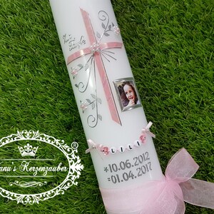 Puede incluir: Una vela blanca decorada con una cruz rosa, detalles florales plateados y una peque&ntilde;a foto enmarcada. La vela tiene el nombre "LILI" escrito con cuentas y las fechas 10.06.2012 y 01.04.2017. Una cinta rosa est&aacute; atada en la base.