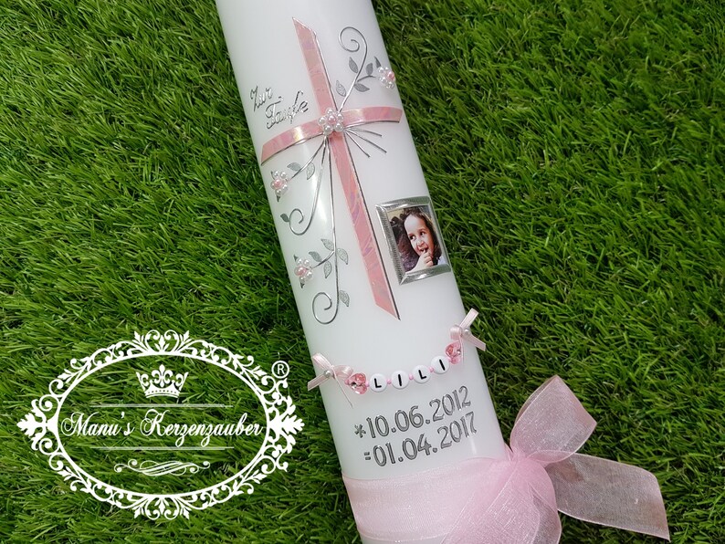 Puede incluir: Una vela blanca decorada con una cruz rosa, detalles florales plateados y una peque&ntilde;a foto enmarcada. La vela tiene el nombre "LILI" escrito con cuentas y las fechas 10.06.2012 y 01.04.2017. Una cinta rosa est&aacute; atada en la base.