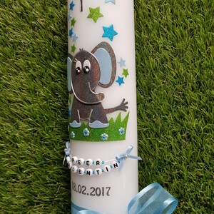Pu&ograve; includere: Candela bianca decorata con un elefante grigio glitterato, stelle blu e verdi e una croce. La candela ha uno striscione con nome di perline e un nastro blu. La data "18.02.2017" &egrave; stampata sulla candela.