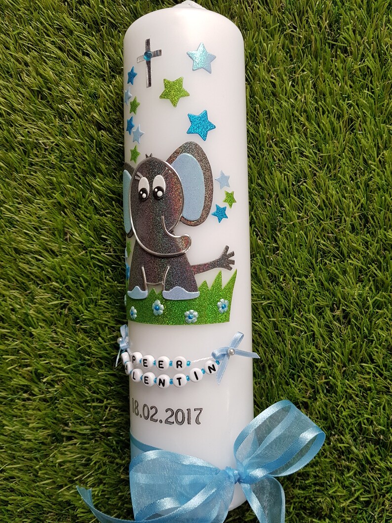 Pu&ograve; includere: Candela bianca decorata con un elefante grigio glitterato, stelle blu e verdi e una croce. La candela ha uno striscione con nome di perline e un nastro blu. La data "18.02.2017" &egrave; stampata sulla candela.