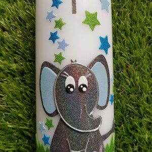 Pu&ograve; includere: Una candela bianca decorata con un elefante grigio glitterato con orecchie e dettagli blu. La candela presenta anche stelle blu e verdi, una croce argentata con una gemma blu e una base verde con fiori blu.