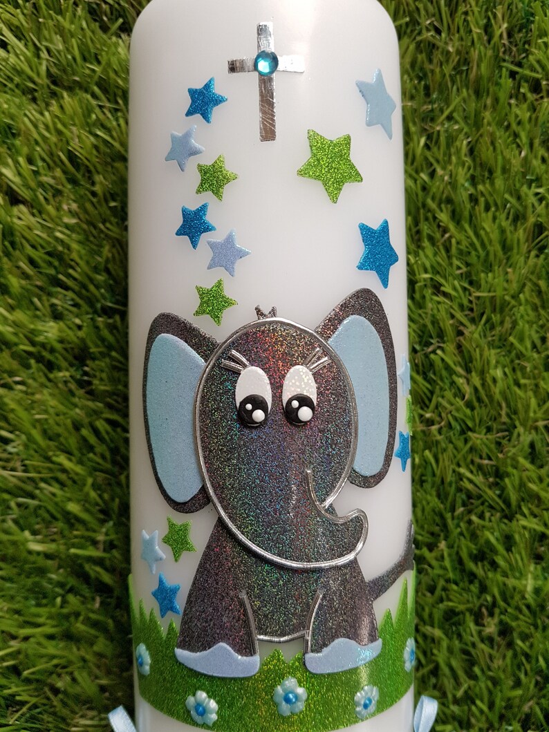 Pu&ograve; includere: Una candela bianca decorata con un elefante grigio glitterato con orecchie e dettagli blu. La candela presenta anche stelle blu e verdi, una croce argentata con una gemma blu e una base verde con fiori blu.
