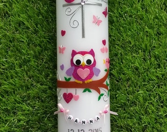 Può includere: Una candela bianca decorata con un gufo rosa, farfalle, cuori e una croce. La candela ha il nome "Hanna" e la data "12.12.2015" stampati. Un nastro e un fiocco rosa decorano la base.