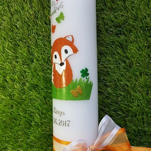 May include: White pillar candle decorated with a cartoon fox, butterflies, and shamrocks. The candle has a green and orange ribbon, and the text "Zur Taufe" and a name and date are printed on the candle.