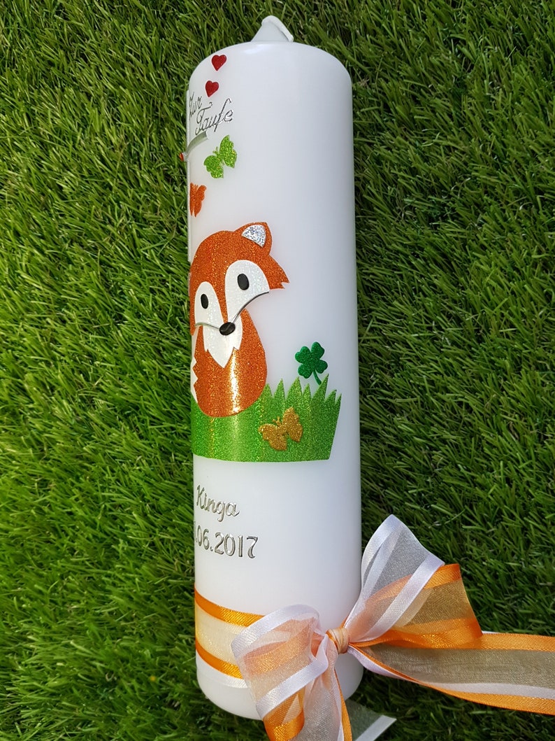 May include: White pillar candle decorated with a cartoon fox, butterflies, and shamrocks. The candle has a green and orange ribbon, and the text "Zur Taufe" and a name and date are printed on the candle.