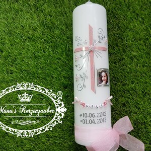 Puede incluir: Una vela blanca decorada con una cruz rosa, detalles florales y un peque&ntilde;o marco de fotos. La vela tiene el nombre "LILI" y fechas impresas. Una cinta y un lazo rosas adornan la base. La vela est&aacute; sobre una superficie verde.