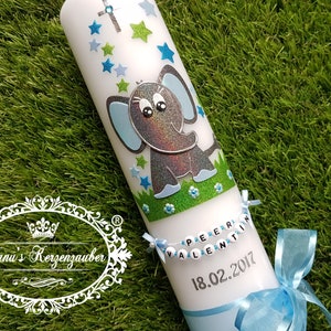Pu&ograve; includere: Una candela bianca decorata con un elefante glitterato, stelle blu e verdi e una croce. La candela ha uno striscione con un nome e una data e un nastro blu. La candela &egrave; su una superficie verde.