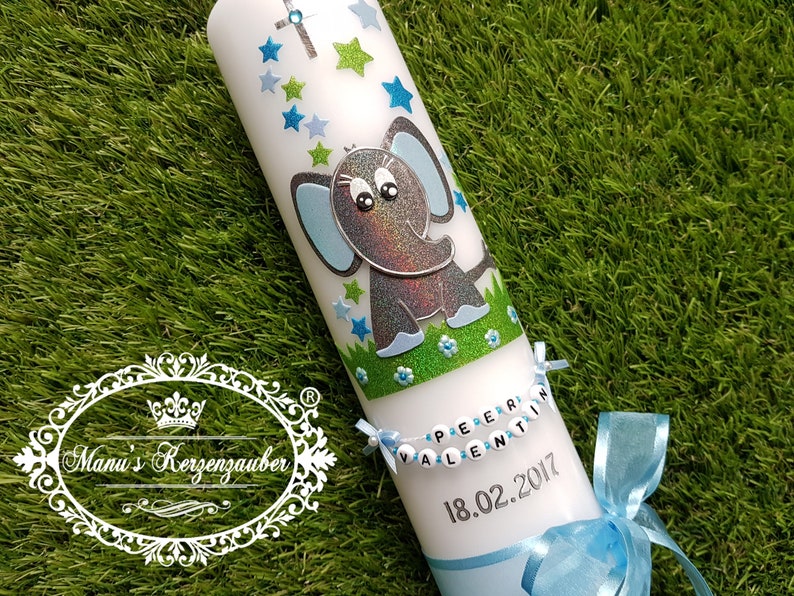 Pu&ograve; includere: Una candela bianca decorata con un elefante glitterato, stelle blu e verdi e una croce. La candela ha uno striscione con un nome e una data e un nastro blu. La candela &egrave; su una superficie verde.