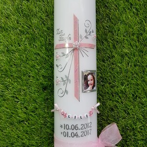 Puede incluir: Una vela blanca decorada con una cruz rosa, detalles florales y una peque&ntilde;a foto enmarcada. La vela tiene el nombre "LILI" escrito con cuentas y las fechas 10.06.2012 y 01.04.2017. Un lazo de cinta rosa adorna la base.