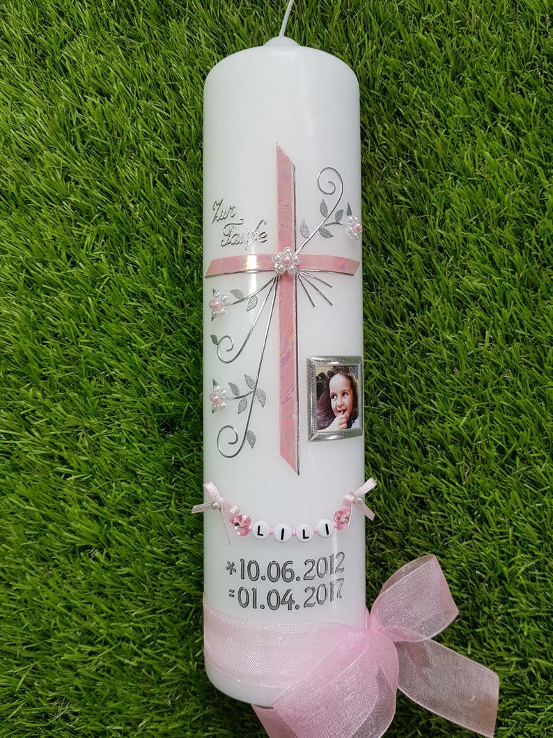 Puede incluir: Una vela blanca decorada con una cruz rosa, detalles florales y una peque&ntilde;a foto enmarcada. La vela tiene el nombre "LILI" escrito con cuentas y las fechas 10.06.2012 y 01.04.2017. Un lazo de cinta rosa adorna la base.