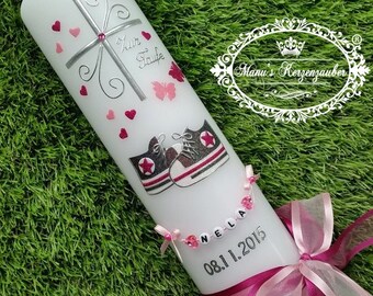 May include: White candle decorated with a silver cross, pink hearts, and butterflies. Features a pair of sneaker illustrations and the name "Nela" with the date "08.1.2015". A pink ribbon and bow complete the design.