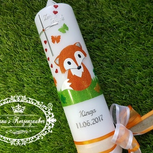 May include: A white candle decorated with a cartoon fox, butterflies, and hearts. The candle has the text "Zur Taufe" and the name "Kinga" with the date "11.06.2017". An orange ribbon and bow adorn the base.