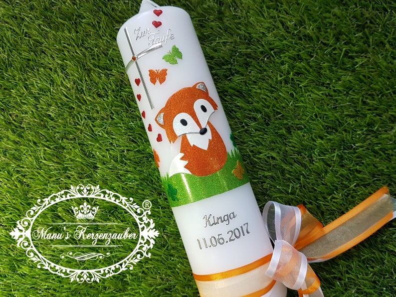 May include: A white candle decorated with a cartoon fox, butterflies, and hearts. The candle has the text "Zur Taufe" and the name "Kinga" with the date "11.06.2017". An orange ribbon and bow adorn the base.