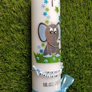 Pu&ograve; includere: Una candela a colonna bianca decorata con un elefante blu e argento, stelle blu e verdi e una croce d'argento. La candela ha un nastro blu e uno striscione con il nome a perline che recita "PEER VALENTIN" e la data "18.02.2017".