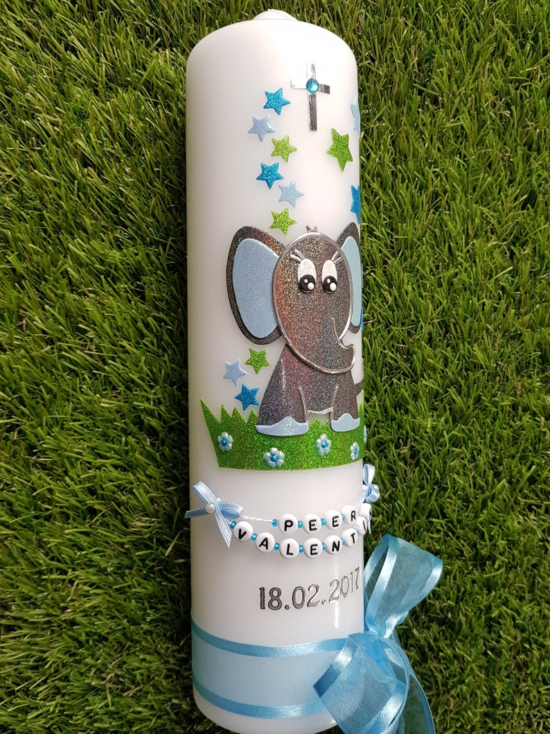 Pu&ograve; includere: Una candela a colonna bianca decorata con un elefante blu e argento, stelle blu e verdi e una croce d'argento. La candela ha un nastro blu e uno striscione con il nome a perline che recita "PEER VALENTIN" e la data "18.02.2017".