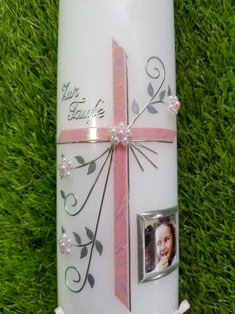 Puede incluir: Vela blanca decorada con una cruz rosa, detalles florales plateados y una peque&ntilde;a foto enmarcada. La vela tiene el texto "Zur Taufe" en plata. La cruz est&aacute; adornada con una flor de perlas.