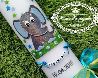 May include: White candle decorated with a glittery gray and blue elephant, blue and green stars, and flowers. The name "JONAS" is spelled out on a string of beads. The date "10.04.2016" is printed below the name. The candle has a blue and green ribbon.