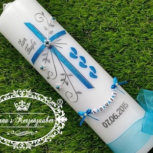 Puede incluir: Vela blanca decorada con una cruz azul, detalles florales y el texto "Zur Taufe". Presenta una cinta azul, el nombre "Louis" en una cadena de cuentas y la fecha 07.06.2015. La vela está sobre una superficie verde.