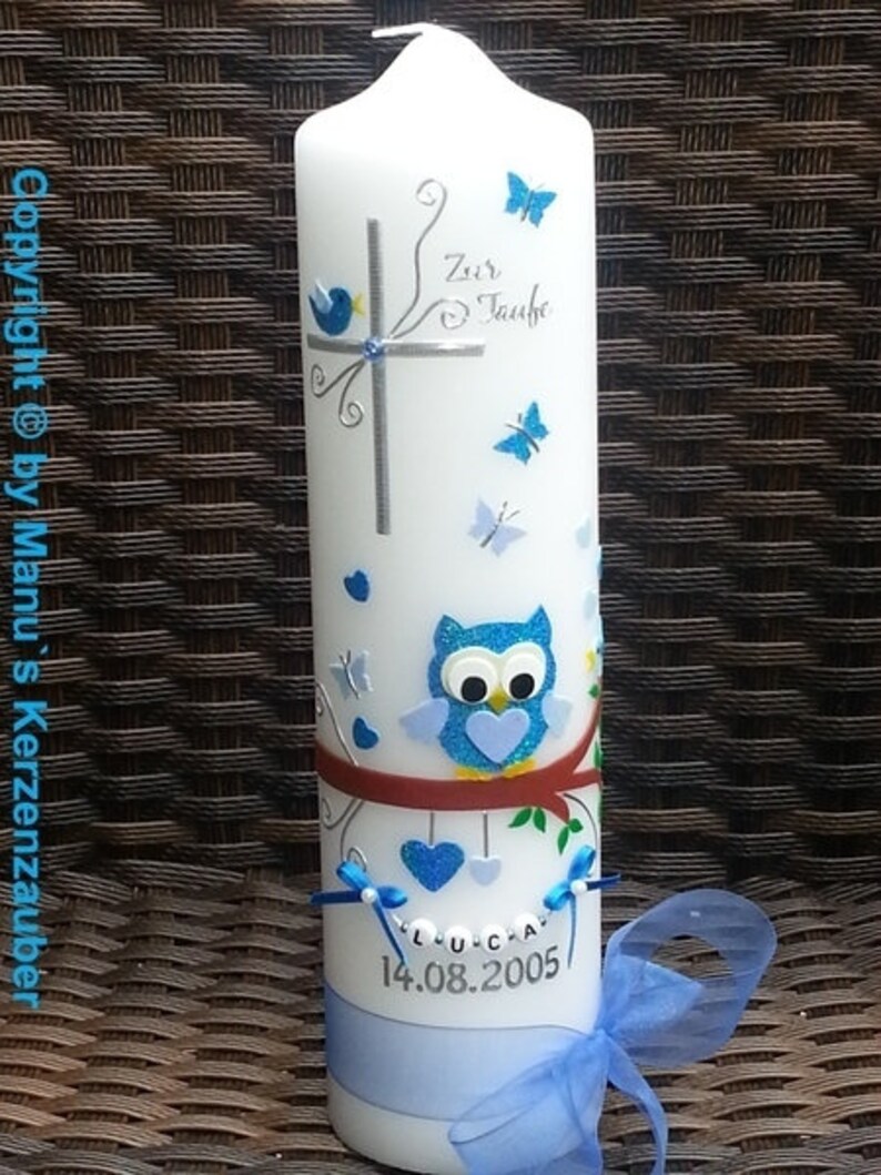 Peut inclure: Une bougie blanche d&eacute;cor&eacute;e de papillons bleus, d'une croix et d'un hibou. La bougie porte l'inscription "Zur Taufe" et le nom "Luca" avec la date "14.08.2005". Un ruban et un n&oelig;ud bleus sont &agrave; la base.