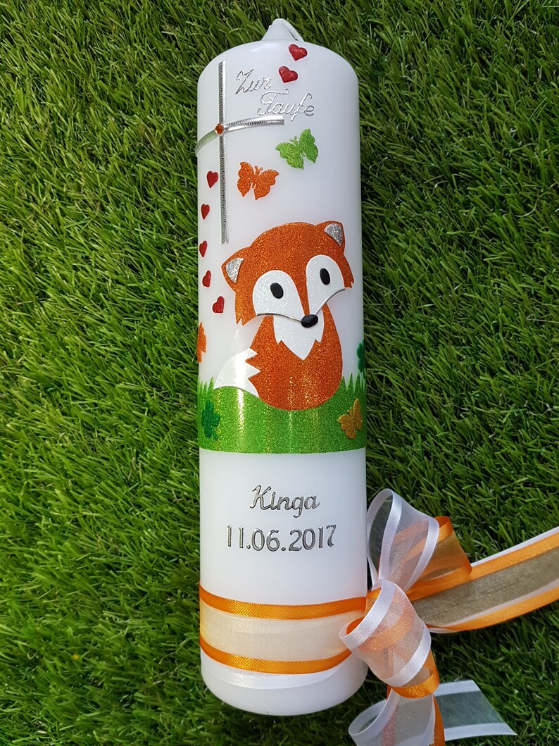 May include: White candle decorated with a glittery orange fox, butterflies, and hearts. The candle has the text "Zur Taufe" and the name "Kinga" with the date "11.06.2017". An orange and white ribbon is tied around the base.