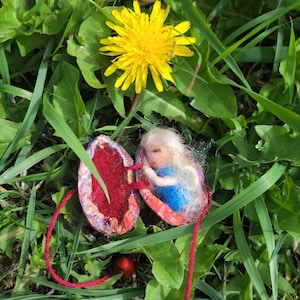Puede incluir: Una pequeña figura de lana fieltrada con largo cabello blanco y cuerpo azul descansa dentro de una cáscara de nuez articulada. El interior está forrado con fibras de color rojo brillante y dos cuerdas rojas con cuentas están unidas al exterior.