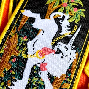 May include: A black and white embroidered patch with a white unicorn and a white dog chained together in a jungle setting. The patch has a yellow border.