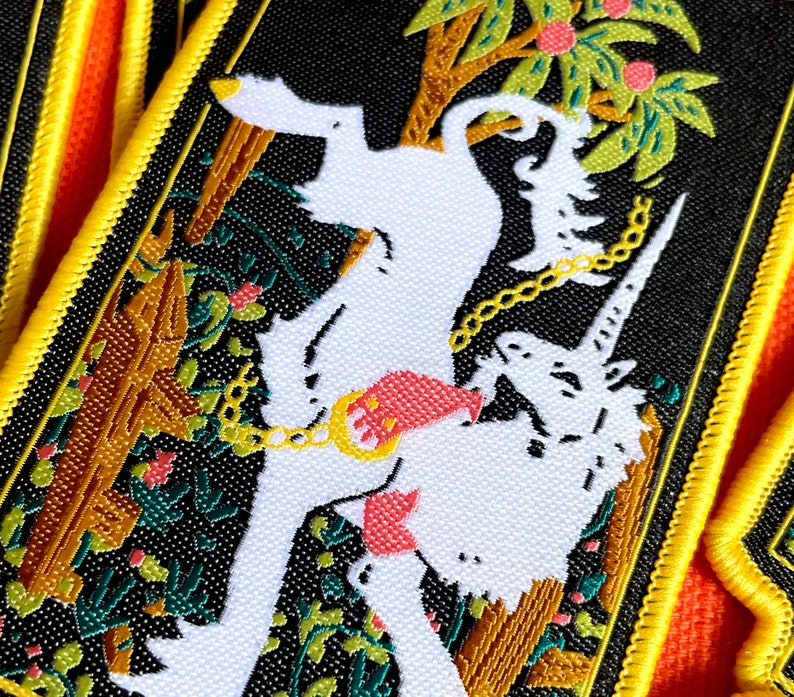 May include: A black and white embroidered patch with a white unicorn and a white dog chained together in a jungle setting. The patch has a yellow border.