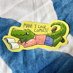 May include: A sticker depicting a green alligator in a pink plaid shirt and blue shorts, relaxing with a book. The text "MAN! I LOVE COMICS!" is above the alligator.