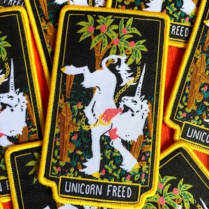 May include: A black and white embroidered patch featuring a white unicorn breaking free from chains. The unicorn is surrounded by green and brown foliage and a tree with orange fruit. The patch has a yellow border and the text "Unicorn Freed" at the bottom.
