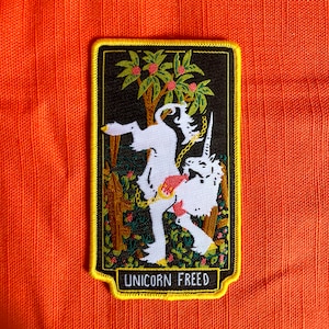May include: A white unicorn with a gold chain is depicted in a black frame with a yellow border. The unicorn is standing on its hind legs in a forest setting with green leaves and red berries. The text "UNICORN FREED" is printed at the bottom of the frame.