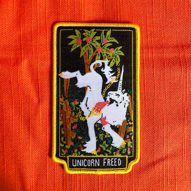 May include: A white unicorn with a gold chain is depicted in a black frame with a yellow border. The unicorn is standing on its hind legs in a forest setting with green leaves and red berries. The text "UNICORN FREED" is printed at the bottom of the frame.