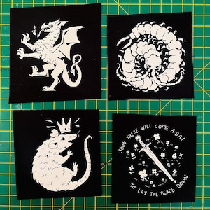 May include: Four square black fabric patches with white screen-printed designs. One features a dragon, one a coiled centipede, one a rat wearing a crown, and one a sword surrounded by flowers with the text, Soon there will come a day to lay the blade down.