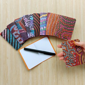 May include: A selection of small notebooks with vibrant, abstract designs, featuring patterns of dots and lines. One notebook is open, displaying blank white pages, with a black pen placed on it. The notebooks are on a light wooden surface.
