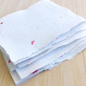 May include: A stack of white handmade paper with colorful flecks and pink triangles. The paper has a rough, textured surface and uneven edges.