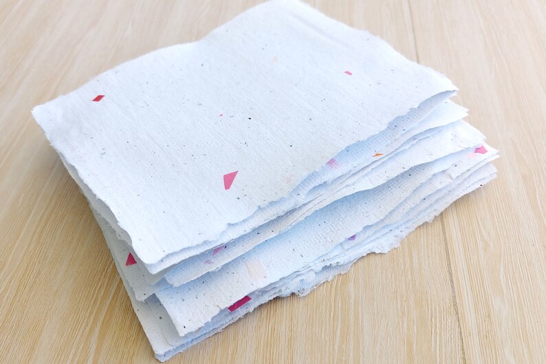 May include: A stack of white handmade paper with colorful flecks and pink triangles. The paper has a rough, textured surface and uneven edges.