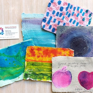 May include: Six handmade paper samples with different textures and colors. The samples are painted with acrylic paint and feature various designs, including dots, stripes, and abstract shapes. The paper is textured and has a rough surface. The samples are arranged on a light wood surface. The text "Textured modeling compound + acrylic paint" is written on one of the samples.