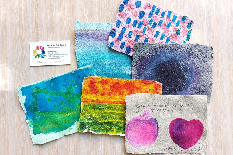 May include: Six handmade paper samples with different textures and colors. The samples are painted with acrylic paint and feature various designs, including dots, stripes, and abstract shapes. The paper is textured and has a rough surface. The samples are arranged on a light wood surface. The text "Textured modeling compound + acrylic paint" is written on one of the samples.