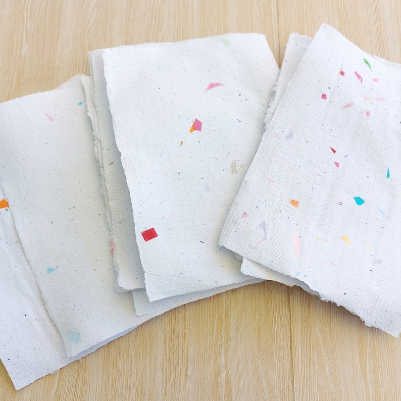May include: A stack of four white sheets of paper with colorful flecks embedded in the paper. The paper has a rough, textured surface.