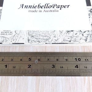 Puede incluir: Primer plano de una regla que mide una pila de papel con una imagen de c&oacute;mic en blanco y negro en la parte superior. La regla est&aacute; marcada en pulgadas y cent&iacute;metros. El papel est&aacute; etiquetado como "Anniebello Paper made in Australia".