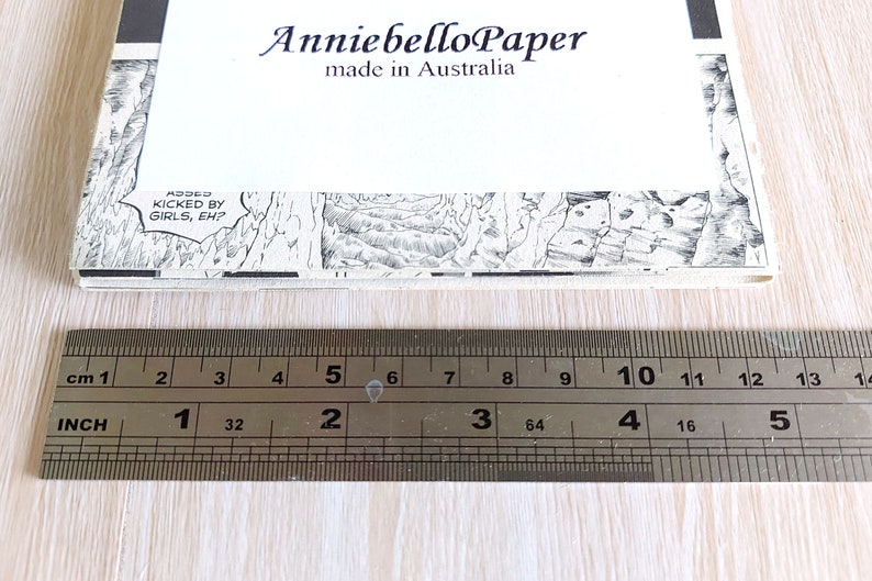 Puede incluir: Primer plano de una regla que mide una pila de papel con una imagen de c&oacute;mic en blanco y negro en la parte superior. La regla est&aacute; marcada en pulgadas y cent&iacute;metros. El papel est&aacute; etiquetado como "Anniebello Paper made in Australia".