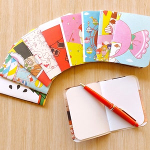 May include: A stack of colourful, illustrated notebooks with a blank notebook and a red pen on top. The notebooks have various designs, including a Ferris wheel, a cartoon character, and a cityscape.