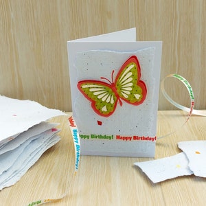 May include: A handmade birthday card with a green and red butterfly on the front. The card is made of white paper with a speckled texture. The card is decorated with a ribbon that says "Happy Birthday!" in colorful letters.