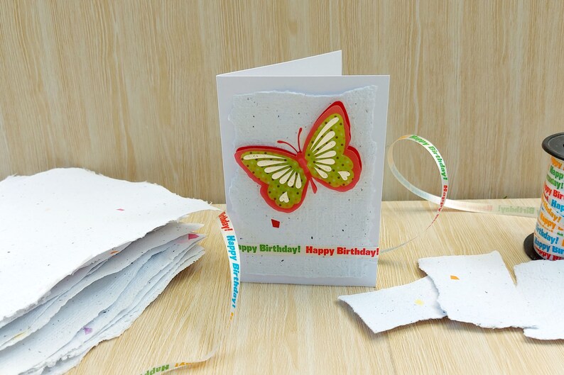 May include: A handmade birthday card with a green and red butterfly on the front. The card is made of white paper with a speckled texture. The card is decorated with a ribbon that says "Happy Birthday!" in colorful letters.