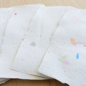 May include: Five sheets of handmade paper with colorful flecks and specks. The paper is white with a rough texture. The flecks are in various colors, including pink, blue, green, and orange.