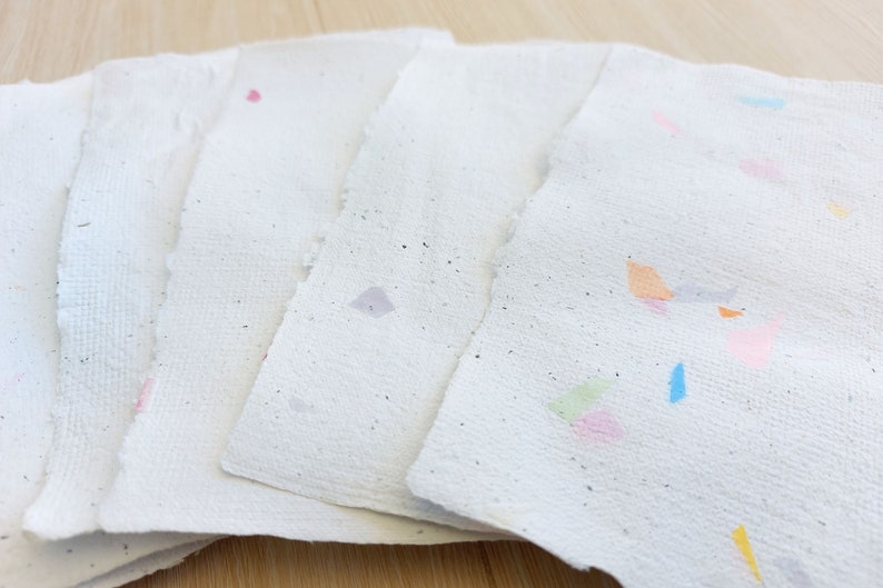 May include: Five sheets of handmade paper with colorful flecks and specks. The paper is white with a rough texture. The flecks are in various colors, including pink, blue, green, and orange.