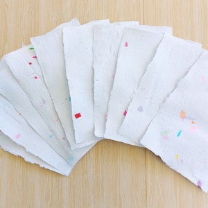 May include: A collection of ten white handmade paper sheets with colorful confetti embedded in the paper. The sheets are arranged in a fan shape.