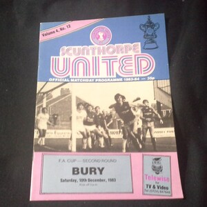 Könnte beinhalten: Ein Vintage-Fußball-Spieltagsprogramm von Scunthorpe United aus der Saison 1983-84. Das Cover zeigt ein Schwarzweißfoto von Spielern in Aktion, mit dem Teamlogo und den Worten "Scunthorpe United" in einer Retro-Schriftart. Das Programm ist für ein FA Cup-Spiel gegen Bury am 10. Dezember 1983.