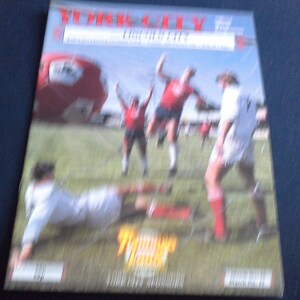 Op de afbeelding: Een rechthoekige tijdschriftomslag met een voetbalwedstrijd in volle gang met spelers in rode en witte tenues op een groen veld. Een grote rode voetbal is zichtbaar in de linkerbovenhoek. De tekst York City staat bovenaan in dikgedrukte letters.